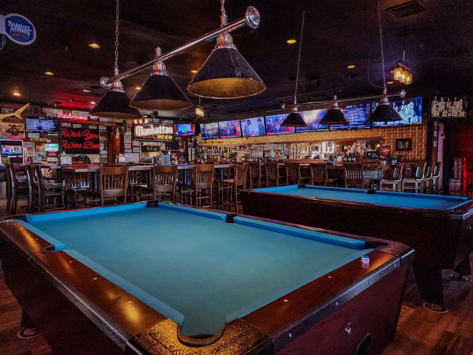 Two pool tables under billiard lamps, the bar and TVs behind